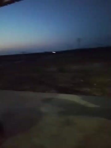 The video shows an outdoor scene at dusk or night. The sky is dark blue. In the distance, there are power lines and utility poles. The ground appears to be dirt with some sparse vegetation and scattered debris. In the foreground, there is a paved surface with shadows. The camera is moving slightly, creating a shaky effect. There are no people or animals clearly visible. The overall impression is of an undeveloped or rural area at low light.