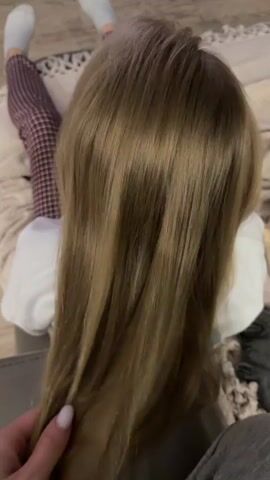 The video shows a close-up view from behind of a person with long, straight, light brown hair. Their hair is being brushed or stroked by a hand. The person is wearing a white shirt and patterned pants. They are lying down on a textured surface that appears to be a blanket or rug, with a white pillow visible behind them. The background is slightly out of focus but suggests an indoor setting.