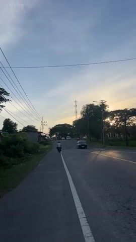 The video shows a road in what appears to be a rural or semi-rural area. The sky is blue with some clouds, and the sun is setting, casting a warm glow on the scene. There are trees and vegetation on either side of the road. A power line runs along the left side of the road. In the distance, there is a tall telecommunications tower. Several vehicles are visible on the road, including cars and a motorcycle. A person is riding a motorcycle on the left side of the road. The road has a white line dividing the lanes. The overall atmosphere is calm and peaceful.