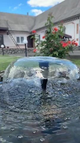A close-up shot of a water fountain in a yard, with a house and rose bushes in the background. The water is clear and bubbling, forming a dome shape. The house appears to be a one-story building with a gray roof and light-colored walls. There are red roses blooming on a bush in front of the house. The sky is blue with some white clouds.