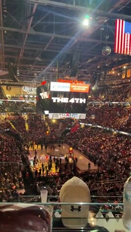 The video shows a high-angle view of a basketball game in Rocket Arena. The Cleveland Cavaliers are playing the Miami Heat; the score at the end of the game is 99-95 in favor of Cleveland. The arena is packed with fans. The jumbotron displays, “The earth is here.” There's a view of drinks and a hat sitting in the foreground. An American flag hangs from the ceiling.