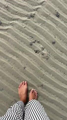 The video shows a top-down view of a person's feet walking on a sandy beach. The sand has ripple patterns and small piles of what appear to be crab mounds. The person is wearing striped pants and a white anklet. The person's feet are bare and are stepping on the sand. The camera is moving forward with the person.