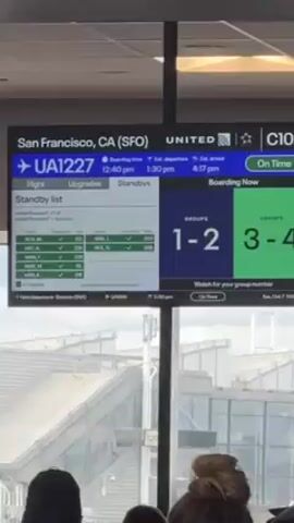 The video shows a crowd of people waiting at an airport gate. An overhead display indicates that United flight UA1227 to San Francisco (SFO) is boarding for groups 1-4, with the flight listed as on time. The display also shows the standby list. Above the crowd is a sign for the "Transfer Shuttle by Gate 71 Terminal A & B." The airport appears well lit with large windows offering a view outside. The people are generally still, waiting for their turn to board.