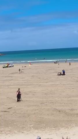 The video shows a sandy beach under a blue sky with scattered clouds. The ocean is visible in the background, with small waves and some people in the water. Several people are on the beach. In the foreground, two people are walking across the sand, one with a dark top and light bottoms, the other in dark clothing. Further up the beach, closer to the water, there are several groups of people. One group is gathered around a white vehicle, possibly a lifeguard vehicle. Another group is sitting or lying on the sand. There are also individuals walking and standing on the beach. A red and yellow flag is planted in the sand. The scene appears to be a typical day at the beach with various recreational activities taking place.