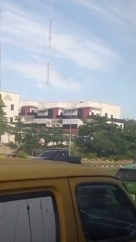 The video shows a view from inside a yellow vehicle, looking out at a modern building with a white facade and dark red accents. Tall communication towers are visible behind the building against a partly cloudy sky. The building appears to be an office or hotel, with a sign that reads "OLISA SUITES". Several cars are driving on a road in front of the building, and trees line the roadside. The interior of the yellow vehicle is visible in the foreground, including the back of a seat and a portion of a window with some markings. The video appears to have been recorded while the vehicle was in motion.