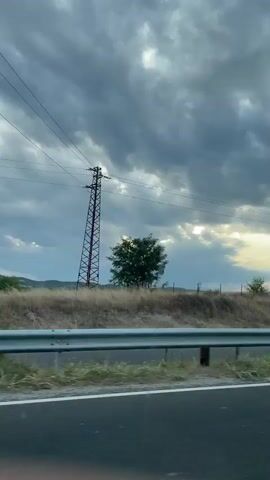The video shows a view from a car moving along a road. The road is in the foreground with a white guardrail. Beyond the guardrail is a dry, grassy field with some scattered bushes and trees. In the distance, there are fence posts and utility poles. The sky is filled with dramatic, dark clouds, with shafts of sunlight breaking through in several places, creating crepuscular rays. A light-colored sedan is visible on the road ahead, moving in the same direction. The overall impression is of a moody, somewhat overcast day, with natural light playing an interesting role.