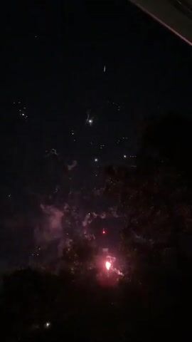 The video shows a fireworks display at night. The sky is dark, with occasional flashes of light from the fireworks. The fireworks are various colors, including red, orange, yellow, and blue. Some of the fireworks explode in bursts of sparks, while others create trails of light across the sky. There are also some clouds visible in the sky, which are illuminated by the fireworks. In the foreground, there are silhouettes of trees. The video is shaky, suggesting it was filmed handheld.