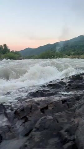 This is a vertical video clip showing a fast-flowing river with white water rapids. The riverbed is composed of dark, jagged rocks in the foreground. In the middle ground, the river churns and splashes, creating a dynamic and powerful visual. The background features a lush green forested hillside and mountains, receding into the distance. The sky is a pale gradient of pink and blue, suggesting either sunrise or sunset. There is a faint plume of smoke visible in the far distance on the right side of the frame.

The scene appears to be a natural landscape, likely in a mountainous or hilly region, given the presence of the mountains and dense vegetation. The weather seems calm, with no indication of rain or strong winds, despite the turbulent water. The time of day is likely early morning or late evening due to the soft lighting and sky colors.

The video captures the raw power and beauty of nature, with the emphasis on the movement and energy of the water. There are no people or man-made objects visible in the clip, focusing solely on the natural environment. The overall mood conveyed is one of natural grandeur and a sense of untamed wilderness. The location context provided indicates this is in Kamennomostsky, Russia.