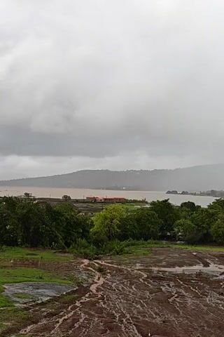 This is a landscape video of a body of water with land on either side. The sky is overcast and appears to be raining. In the foreground, there is a muddy, uneven terrain with some patches of green grass. Further back, there are trees and some buildings near the water. The water is brown, indicating it may be murky or carrying sediment. The overall scene suggests a natural environment, possibly after or during heavy rainfall.