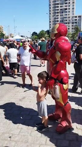The video shows a young boy and a person in an Iron Man costume standing on a brick-paved area with crowds of people in the background. The boy is shirtless, wearing gray shorts and dark shoes, and is facing forward with his hand near his mouth. The Iron Man costume is red and gold and is standing to the right of the boy, with one arm extended. The background includes a street with pedestrians, a tall building, trees, and a clear blue sky. The scene appears to be taking place during a daytime event in a public area.