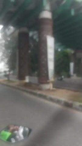 The video shows a first-person perspective of someone riding a motorcycle. The rider is wearing a green jacket with a large white circular logo. The motorcycle has a side mirror, which reflects the rider and the green jacket. The road appears to be paved and has a white line marking the edge. In the background, there are trees, buildings, and streetlights. The video is blurry and appears to be taken while in motion.