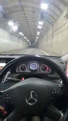 A first-person perspective video captures the interior of a car driving through a tunnel. The black steering wheel with a prominent Mercedes-Benz logo is in the foreground, framing the dashboard. The dashboard features analog gauges for speed, RPM, fuel, and temperature, along with a digital display showing "81 km/h" in the center. An infotainment screen is visible to the left, and a smartphone is mounted to the right of the dashboard. The car is traveling through a well-lit tunnel with concrete walls and numerous bright lights illuminating the ceiling. Some water droplets or reflections are visible on the windshield. The road ahead appears clear, with no other vehicles immediately in view. The driver's hands are visible on the steering wheel, indicating active driving through the tunnel.