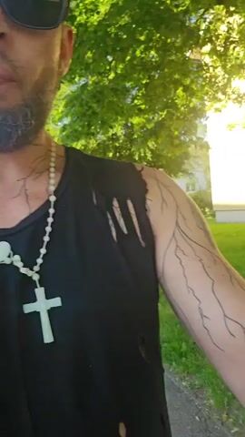 A medium shot focuses on a person from the chest up, facing left. They are wearing a black, ripped tank top and a beaded necklace with a white cross. The person has a beard and sunglasses. Their left arm is visible, with a tattoo of what appears to be a tree or lightning branches extending from the shoulder down the arm. The background shows green trees, a building, and a sidewalk. The lighting suggests it is daytime and sunny. The video pans slightly to the right, revealing more of the tattooed arm, which has a large tattoo of headphones with a green circular logo that reads "genge" backwards. The person's right arm is also partially visible.