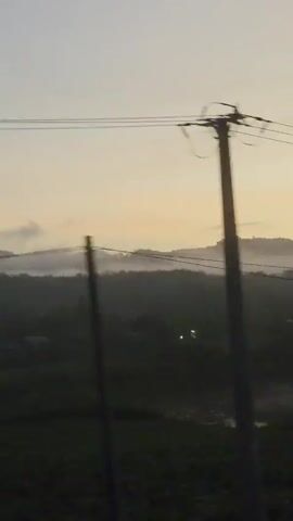 The video shows a landscape at dawn or dusk, with a hazy or misty atmosphere obscuring the details of the distant hills. Power lines are visible in the foreground, running horizontally across the upper part of the frame. Below, the land appears as a dark, undifferentiated mass, likely fields or vegetation, with a few scattered light sources. The sky is a pale, washed-out color, hinting at the transition between day and night. The camera appears to be moving, as the perspective shifts slightly during the video.