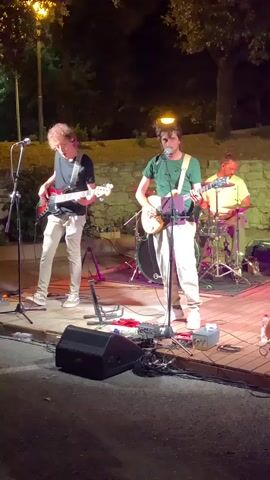 A band is performing music outdoors at night. The band consists of at least three musicians. The person in the foreground on the left is playing a bass guitar and singing into a microphone. The person in the center is playing an electric guitar and singing into a microphone. The person on the right is playing drums. There are instruments and equipment on a raised wooden stage. The background appears to be a park or outdoor area with trees and a stone wall. The lighting is dim, with spotlights illuminating the musicians. The overall scene suggests a live music performance in an informal setting.