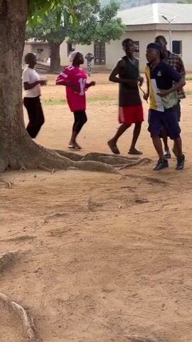 In Tabu, Nigeria, a group of approximately seven to eight young people are seen running and jogging in a loose circular path around a large tree with prominent exposed roots. The setting is an unpaved, reddish-brown dirt ground with several buildings visible in the background, suggesting a community or village. The time of day is bright and sunny.

The individuals are dressed in casual attire, including t-shirts, shorts, and a plaid shirt. One person wears a bright pink jersey with the number "10" clearly visible on the back. Another person, wearing a dark blue top and shorts, carries a green cloth or plastic bag, while a person in the pink jersey holds a drink. Some participants are wearing sandals, others are in shoes, and at least one appears to be barefoot. The activity seems energetic, possibly a form of group exercise, game, or rhythmic running.