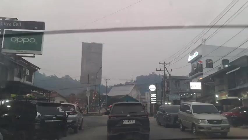 The video shows a street scene with several cars and motorcycles. The sky is overcast and there are buildings on either side of the street. There is a tall billboard in the distance. The camera appears to be mounted inside a vehicle.