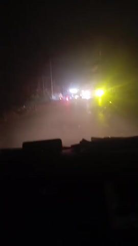 The video shows a nighttime drive on a paved road. The view is from inside the vehicle, looking out through the windshield. Headlights from oncoming vehicles illuminate the road ahead. There are also motorcycles with lights on. The road appears slightly wet. The dashboard of the vehicle is visible in the foreground, largely obscured by darkness. The environment seems to be a rural or semi-rural area with some trees or vegetation visible on the sides of the road.