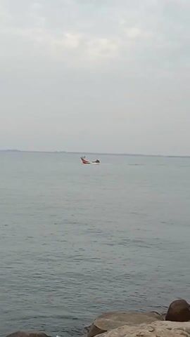 The video shows a distant view of two boats moving across calm water. The boats appear small, with one being orange and white and the other yellow. The sky is overcast, and the horizon is slightly visible in the distance. In the foreground, there are a few rocks. The overall scene suggests a calm day on the water.