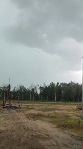 The media file depicts an outdoor scene, likely in an industrial or resource extraction area. The primary subjects are oil pumpjacks, industrial equipment, and a wooded landscape under a cloudy sky. The scene appears to be a barren, cleared area with dirt paths, suggesting an active or recently developed site.

In the foreground, a dirt road with tire tracks indicates vehicle movement. The middle ground features several oil pumpjacks, characterized by their distinctive up-and-down pumping motion. One pumpjack is particularly prominent on the left side of the frame, showing its metallic structure and red accents. Other smaller structures and pipes are also visible around the pumpjacks, consistent with an oil field operation.

The background is dominated by a dense line of trees, forming a backdrop to the industrial activity. The trees are mostly deciduous, and some appear to be bare or sparsely leaved, suggesting it might be late autumn, winter, or early spring. A tall, slender structure, possibly a communication tower or another piece of industrial infrastructure, stands to the far right.

The sky is heavily overcast with dark, heavy clouds, creating a somber and potentially ominous atmosphere. The lighting is diffused, and there are no strong shadows, indicating a lack of direct sunlight, typical of a heavily clouded day or possibly approaching bad weather.

The activity taking place is the operation of oil pumpjacks, a process of extracting crude oil from underground reservoirs. This implies an ongoing industrial operation at this location.

There is no visible text in the media file. The overall emotion conveyed by the scene is one of industrial endeavor set against a natural, yet somewhat bleak, landscape under a threatening sky. The location is identified as Ровенскаслабодскі сельскі Савет, Belarus, which provides geographical context for the scene.