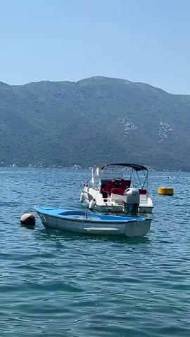 The video shows two boats on a body of water with a mountainous backdrop. In the foreground, a small, white rowboat with a blue rim is tethered to a brown buoy. Behind it, a larger white motorboat with a black canopy and red seats is anchored, its silver outboard motor visible. The water is calm and blue. The mountains in the background are covered in greenery. A yellow buoy is visible in the distance. The sky is clear and blue.