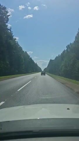 A first-person perspective shot captures a car driving on a highway surrounded by dense trees. The sky is a clear blue with scattered white clouds. A single dark-colored car is visible ahead on the road, traveling in the same direction. The road has clear lane markings, with a white line dividing the lanes. The sun appears to be shining, casting some light and shadow on the scene. The shot is focused on the road and the scenery, with the dashboard of the filming vehicle visible at the bottom of the frame.