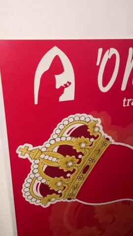 The video shows a close-up, panning shot of a red sign with white text and graphics. The text appears to read "'O Muro, trattoria e pizzeria" on the left side and "aciellello" on the right. Below the text, there is a graphic of a golden crown with red jewels, and further to the right, a graphic of a two-headed bird with yellow beaks and red bodies. In the background, a shelf with several bottles of rose wine is visible. The sign appears to be displayed on a wall.