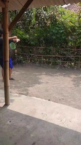 A person is playing badminton outdoors. They are wearing a black t-shirt and blue shorts. The person is standing under a bamboo-like structure with a corrugated roof. They are holding a badminton racket and appear to be about to hit a shuttlecock. The ground is dirt and gravel, and there is greenery in the background, including trees and a fence. The lighting suggests it is daytime.