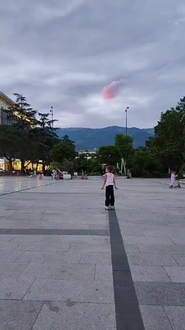 A young girl on roller skates stands in a paved plaza, her body angled to the left. She is wearing a pink t-shirt and black pants. Behind her, a single pink cloud is visible in a twilight sky, contrasting with the muted grays and purples of other clouds. In the background, trees and mountains can be seen. Other people are seated in the background on benches. The overall scene appears to be a public space at dusk.