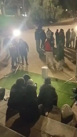The video shows a group of people gathered in an outdoor space at night. The area is lit by a standing light. Some people are standing and interacting in the center, while others are seated on steps, some looking at their phones. The ground is a mix of dirt and artificial turf. A white container is also visible. The scene appears to be a casual gathering, possibly a filming session or a relaxed social event.