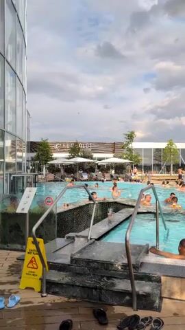 The video shows an outdoor swimming pool with many people in it. The pool is surrounded by a deck area and a building with large windows. There are trees and umbrellas around the pool. The sky is cloudy. A warning sign is visible near the stairs leading into the pool. People are swimming, talking, and relaxing in the water.