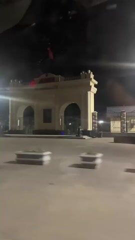 A nighttime video captures a vehicle's perspective moving past a large, ornate gateway. The gateway is illuminated by bright lights, with text visible above the arches. The area in front of the gateway appears to be a paved plaza with planter boxes containing greenery. To the left of the gateway, a long wall with colorful murals is visible. Beyond the gateway, trees and more lights are faintly discernible in the darkness. The video shows motion blur due to the movement of the vehicle.