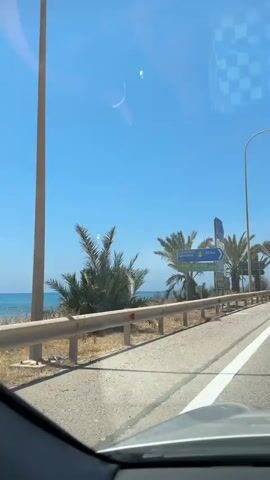 A car is driving down a paved road bordered by a guardrail. On the other side of the guardrail, there is a dry, dusty embankment with sparse vegetation and several palm trees. Beyond the embankment, the bright blue sea is visible. The sky is clear and blue with a few faint reflections on the car's windshield. Several road signs are mounted on a pole near the beginning of the video, indicating distances to various locations. The weather appears to be sunny and warm.