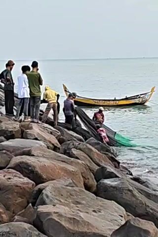 Several individuals are gathered on a rocky shoreline, engaged in what appears to be fishing activities. Six figures stand atop a collection of large rocks, observing or participating in the task at hand. Closer to the water's edge, four individuals are actively handling a dark, possibly green, fishing net that extends into the water. A small, yellow boat is anchored in the sea a short distance away. The environment includes a wide expanse of water extending to the horizon under an overcast sky. The foreground is dominated by the irregularly shaped rocks that make up the shoreline.