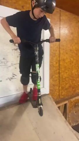 A young male, wearing a helmet, a dark t-shirt, and black shorts, rides a scooter in an indoor skatepark. The skatepark features wooden ramps and platforms of varying sizes. The camera pans to follow the rider as he navigates the park, at one point ascending a ramp and approaching a large black surface. The lighting is artificial, suggesting an indoor environment.