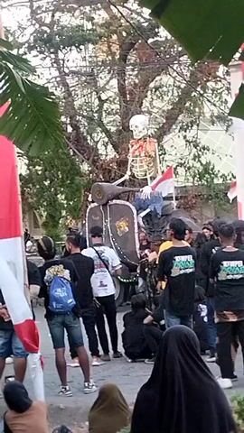 A skeleton is mounted on a large, dark-colored scooter, positioned in the center of a street or public gathering. The skeleton is wearing blue pants and a shirt. A large Indonesian flag is prominently displayed on a pole in the foreground to the left, and a smaller Indonesian flag is visible behind the skeleton on the scooter. A crowd of people, many wearing dark t-shirts, are gathered around the scooter and skeleton, observing the display. Some individuals are standing, while others are seated on the ground. The background shows trees and buildings, suggesting an outdoor urban or semi-urban environment. The overall scene appears to be a parade, festival, or organized event.