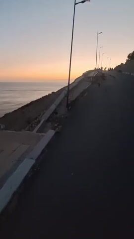 The video captures a scenic view of the coastline at dusk, with the sun setting over the ocean. The camera is positioned on an asphalt road that appears to be damaged with visible cracks and erosion along the edge, bordering a steep embankment leading down to the sea. A concrete retaining wall along the embankment is also showing signs of damage and collapse. Several lampposts line the side of the road, illuminating the path. In the distance, on the higher part of the embankment, a group of people can be seen gathered, possibly observing the sunset or enjoying the view. The sky displays a gradient of warm colors, transitioning from orange and yellow near the horizon to a soft blue overhead. The overall atmosphere is serene and picturesque, despite the apparent damage to the road and infrastructure. The time of day is clearly evident as sunset, indicated by the warm hues in the sky and the diminishing light. The location appears to be a coastal area, likely a road overlooking the ocean, possibly in Safi, Morocco, given the context. The mood evoked is one of natural beauty and tranquility, with a hint of melancholy due to the visible signs of decay and potential danger. There is no visible text in the video.