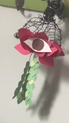 A close-up, static shot shows a wall decoration. The decoration consists of a black wire sculpture with red paper flower petals and a white paper eye with a brown iris in the center. Green paper leaves hang from the flower. A shadow of the decoration is visible on the white wall. A small metal hook is visible in the upper left corner. The background includes part of a green wall hanging.