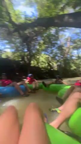 The video shows three people in inner tubes floating down a river in a lush, green environment. They are all wearing life jackets and helmets. The person filming is in a bright green inner tube and is mostly out of focus. The other two people, one in a green inner tube and the other in a blue one, are more visible. The person in the blue inner tube is face down in the water at one point, but then they sit up. The river is surrounded by trees, creating a dense forest canopy overhead. The sunlight filters through the leaves, creating dappled light on the water. The overall atmosphere is one of relaxation and enjoyment.