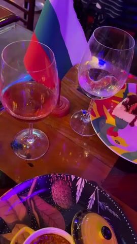 A video displays a close-up of a dining table in a dimly lit setting illuminated with purple and blue light. Two wine glasses are visible; the left one contains a reddish liquid, and the right one holds a clear liquid. Between the glasses, a small Russian flag on a wooden stand is positioned. To the right, a colorful plate with a stylized rooster design holds a slice of cheesecake topped with chocolate sauce and sliced strawberries. A partially visible dark-patterned plate with a small yellow bowl containing food is in the bottom left. A person's hand holding a mobile phone is briefly seen in the background. The camera pans from a general view of the table to a close-up of the cheesecake.