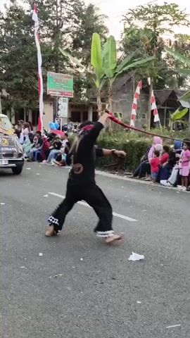 A male performer in black clothing and a red headband is seen performing a martial art or dance routine outdoors on a paved road. He is barefoot and wears a black shirt with a white emblem on the left chest and black pants with a checkered pattern at the calves. He holds a red whip or rope in his right hand and a flat, dark object, possibly a shield or another prop, in his left hand. He is captured mid-movement, with his body slightly twisted and his arms extended. The background shows a crowd of spectators sitting on chairs and along the roadside, with some people standing. There are Indonesian flags (red and white) mounted on poles, palm trees, and other foliage visible. The setting appears to be a parade or a local festival, with a truck partially visible on the left side of the frame. The ground is littered with some paper debris.