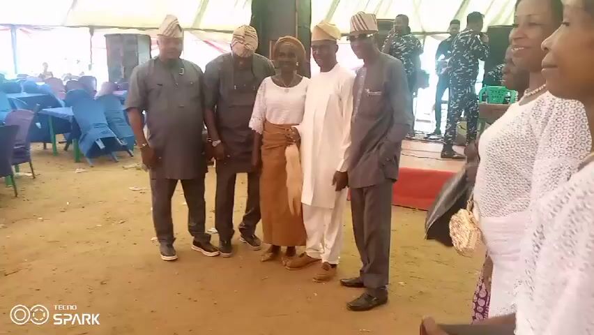 The video shows a group of people standing together, likely for a photograph. They are dressed in a mix of traditional and formal attire. All the men are wearing traditional Yoruba hats, and some are dressed in suits. The woman in the center is wearing a white top and a brown wrapper. Behind them is a large tented area with tables and chairs, suggesting this is an event or celebration. There are uniformed officers in the background, indicating a formal or public gathering.  The ground is a sandy color. A watermark indicates that the video was taken with a Tecno Spark phone.