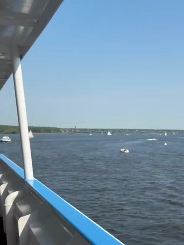 The video shows a body of water with several boats, some with sails, scattered across it. The water appears calm with small ripples. In the distance, a tree-lined shore is visible. The sky is clear and blue. The video appears to be filmed from a boat, as indicated by the railing and part of the boat's structure in the foreground. There are no people clearly visible in the water or on the shore. The overall impression is of a leisurely day on the water.