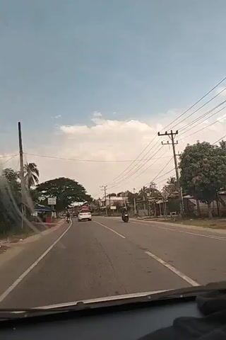 The video shows a view from inside a vehicle driving on a road. The road has two lanes and is lined with power lines, trees, and buildings. There are several other vehicles on the road, including cars, motorcycles, and trucks. One of the trucks is a tanker truck with the logo of Pertamina, an Indonesian oil and gas company. The sky is blue with white clouds.