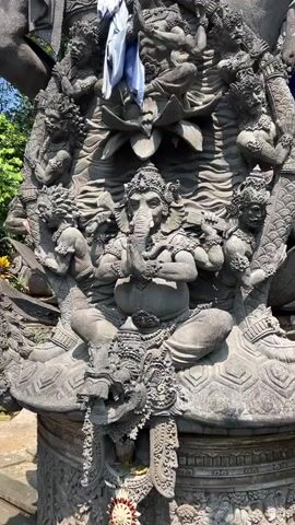 A stone statue depicts a multi-armed deity with an elephant head, seated in the center. The deity's hands are clasped together in front of its chest. Surrounding the central figure are smaller figures carved into the stone, many appearing to be human-like with elaborate headdresses and jewelry. Some of these figures are positioned on the sides of the main deity, while others are situated below it. The statue itself is intricately detailed, with patterns and textures carved into the stone. The overall structure appears to be part of a larger monument or temple. In the background, foliage and a bright blue sky are visible, suggesting an outdoor setting. There are pieces of white and blue cloth draped over parts of the statue, possibly offerings or decorations. The lighting suggests it is daytime.