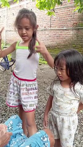 Two young girls stand outside in a yard covered in small gravel. The girl on the left is slightly older, with her dark hair in a partial ponytail. She wears a white tank top with green trim and patterned shorts. She makes a peace sign near her face. The girl on the right, with dark hair that hangs straight down, wears matching pajama-style shorts and top with a light pattern. She puts something in her mouth, likely a piece of food being offered by a person seated out of frame at the bottom of the shot, whose blue patterned clothing is visible. Behind the girls, there is a brick wall partially covered in moss. A blue bicycle sits to the left, and green foliage hangs above.