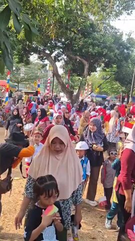 This is a vertical video of an outdoor gathering, likely a festival or celebration, with many people, including children dressed in costumes. The environment appears to be a street or open area lined with trees and some buildings. The crowd is dense, and many individuals are wearing red and white attire, possibly related to a national holiday. Children are dressed in various costumes, including what appear to be police uniforms, traditional outfits, and some carrying small drums. Adults are also present, some holding flags or decorations. The overall atmosphere seems festive and crowded, with a focus on children's participation.