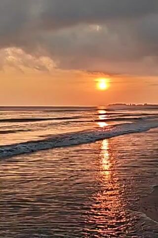 The video displays a scenic ocean beach at sunset, with the sun positioned above the horizon reflecting brightly on the water. Gentle waves roll onto the sandy beach, their crests tinged with white foam. Dark clouds hover in the sky above, contrasting with the orange and golden hues of the sunset. In the distance, a faint outline of land is visible.