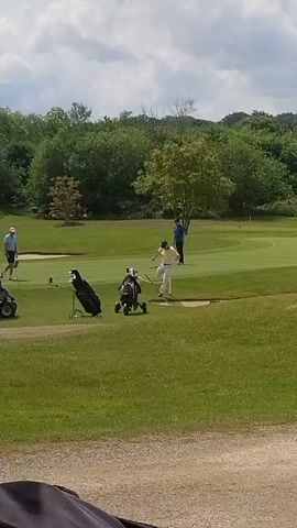 The scene is a golf course on a sunny day. Four golfers are visible. One is wearing a light blue shirt and white hat, another a yellow shirt and white pants, and the third a blue shirt. The fourth golfer is partially obscured. A golf cart is parked on the left side of the frame. Two golf bags are positioned near the golfers. The background features green trees and a cloudy sky. The video appears to be taken from inside a vehicle, with part of the vehicle's interior visible in the lower left corner.