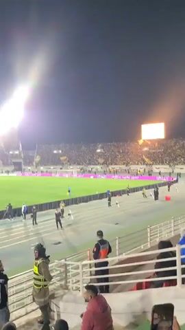 The video shows a stadium at night filled with people in the stands, with a large video screen visible at one end. There are security personnel and other people on the track surrounding the soccer field. Some people in the foreground appear to be watching the game from the stands. A person is holding up a phone to show a video call. A stadium floodlight is on.